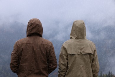 Photo of Man and woman in raincoats enjoying mountain landscape under rain, back view