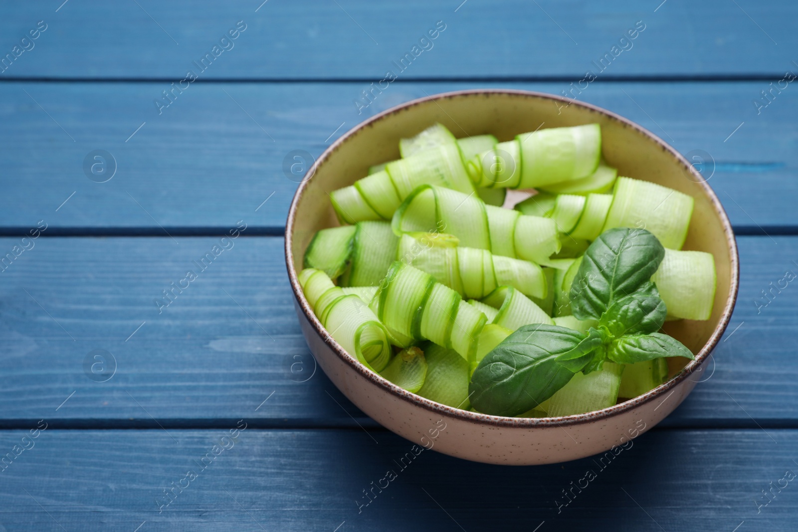Photo of Slices of fresh ripe cucumber and basil in bowl on blue wooden table, space for text