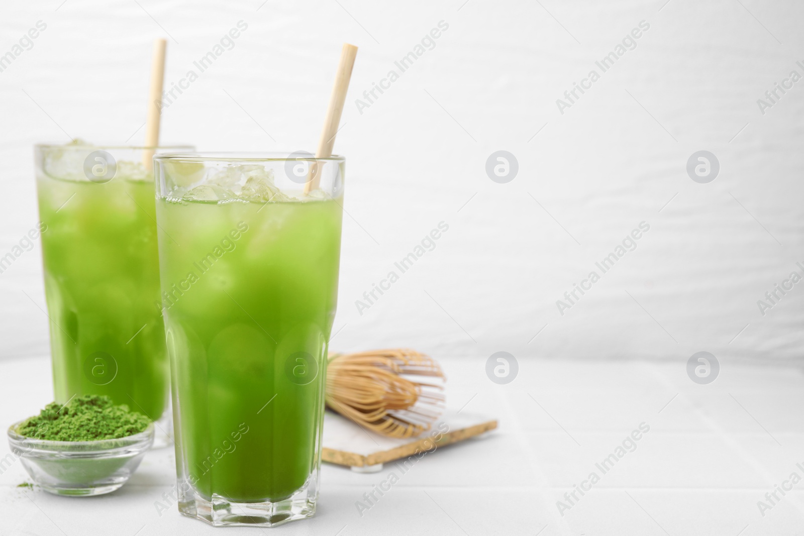 Photo of Delicious iced green matcha tea, powder and bamboo whisk on white tiled table, space for text