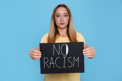 Young woman holding sign with phrase No Racism against light blue background, focus on hands
