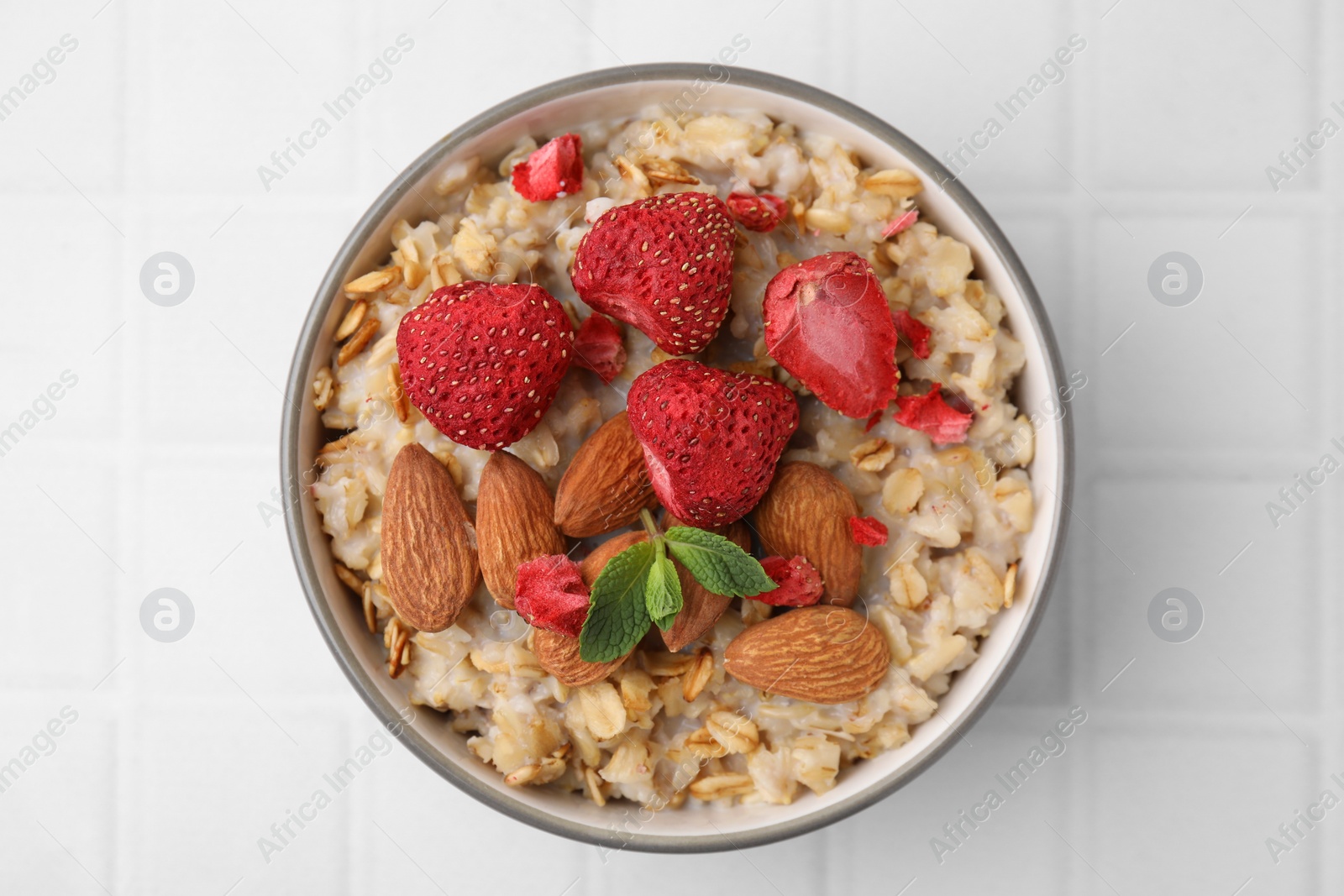 Photo of Delicious oatmeal with freeze dried strawberries, almonds and mint on white tiled table, top view