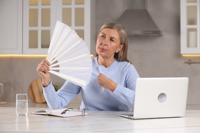 Photo of Menopause. Woman waving hand fan to cool herself during hot flash at table in kitchen