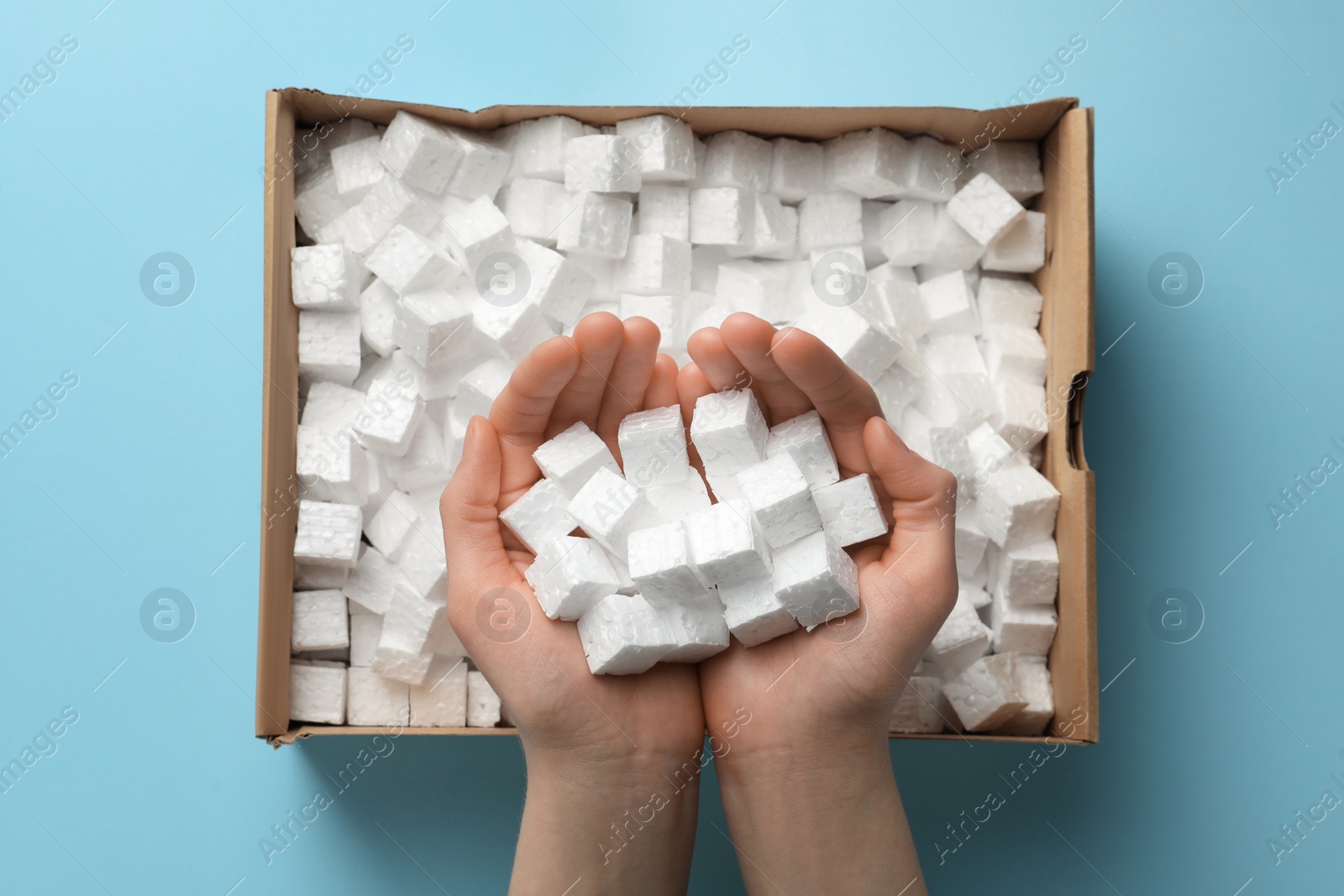 Photo of Woman holding heap of styrofoam cubes over box on light blue background, top view
