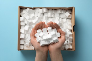 Woman holding heap of styrofoam cubes over box on light blue background, top view