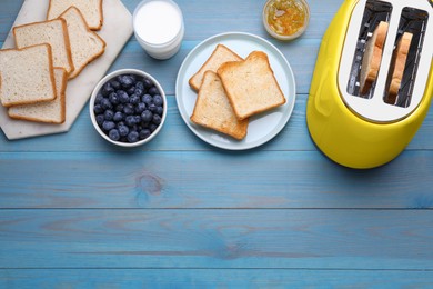 Photo of Yellow toaster with roasted bread, glass of milk, blueberries and jam on light blue wooden table, flat lay. Space for text