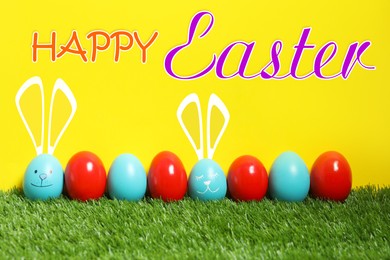 Image of Happy Easter. Two eggs with drawn faces and ears as bunnies among others on green grass against yellow background