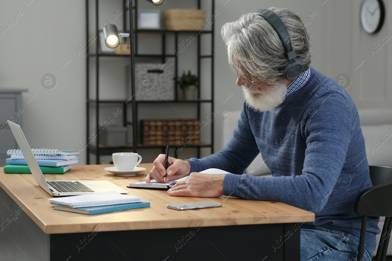 Photo of Middle aged man with laptop, notebook and headphones learning at table indoors