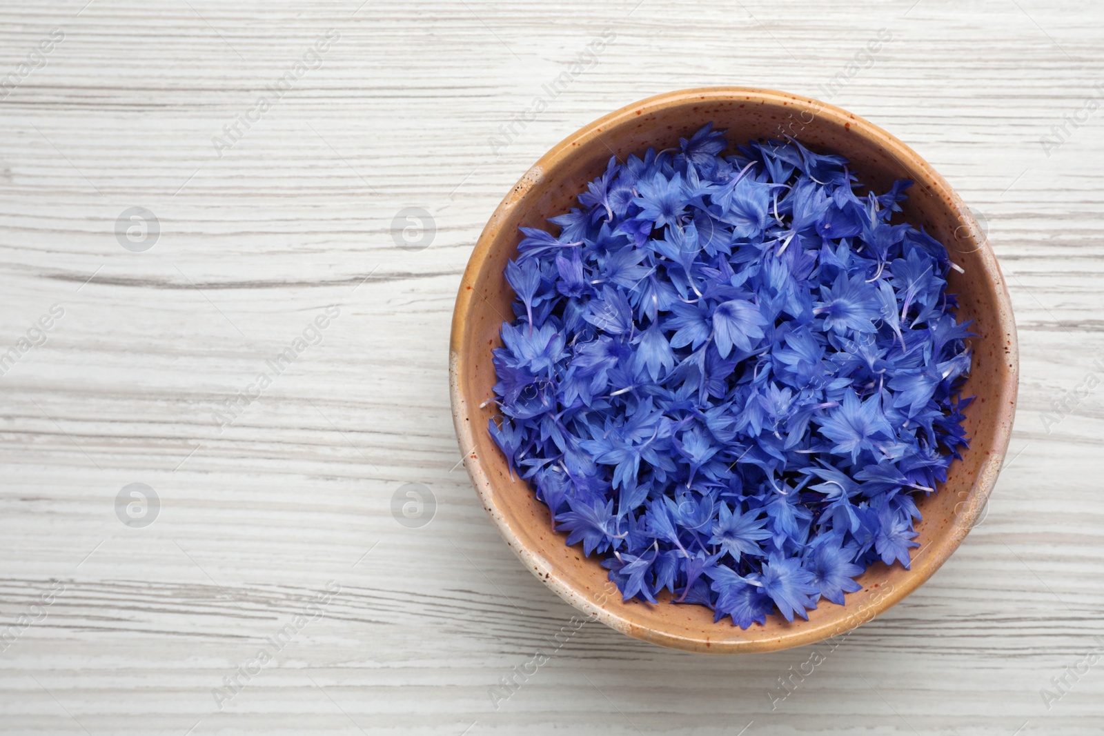 Photo of Beautiful blue cornflowers petals in bowl on white wooden table, top view. Space for text