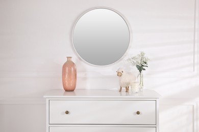 Trendy round mirror and chest of drawers with accessories near white wall. Interior element
