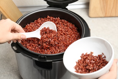 Photo of Woman putting brown rice into bowl from multi cooker in kitchen, closeup