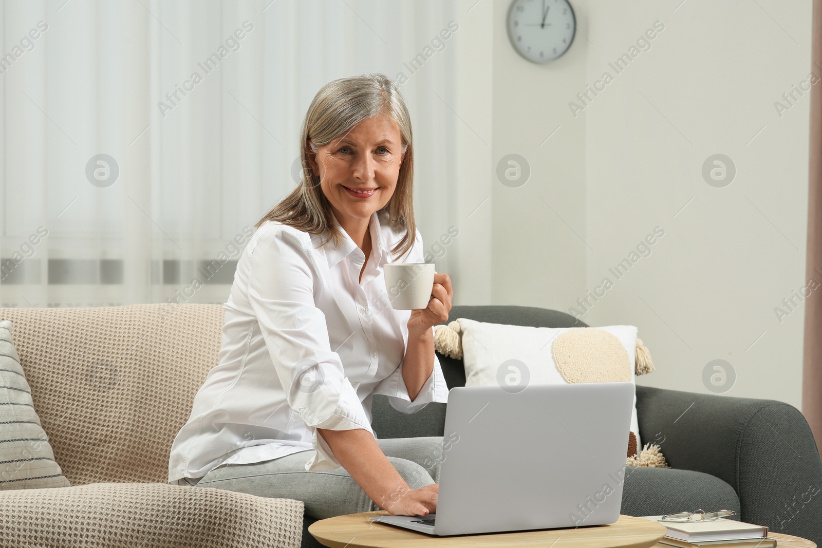 Photo of Beautiful senior woman with cup of drink using laptop at home