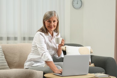 Beautiful senior woman with cup of drink using laptop at home