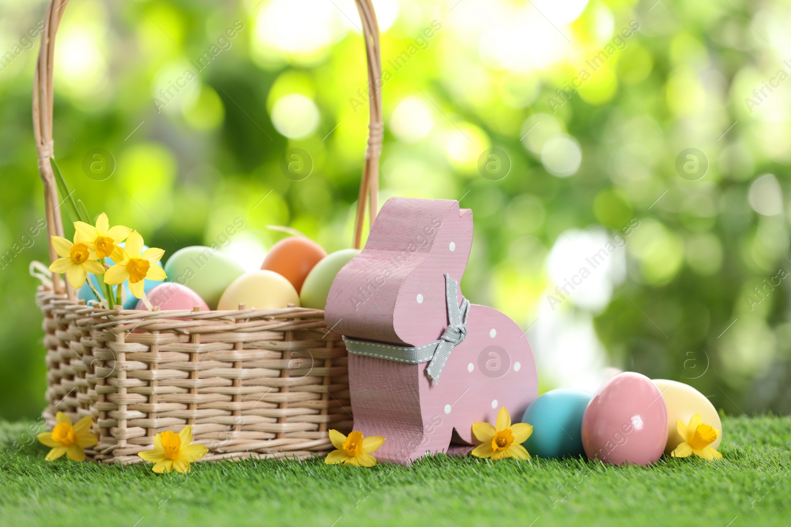 Photo of Cute wooden Easter bunny with wicker basket and dyed eggs on green grass against blurred background, space for text