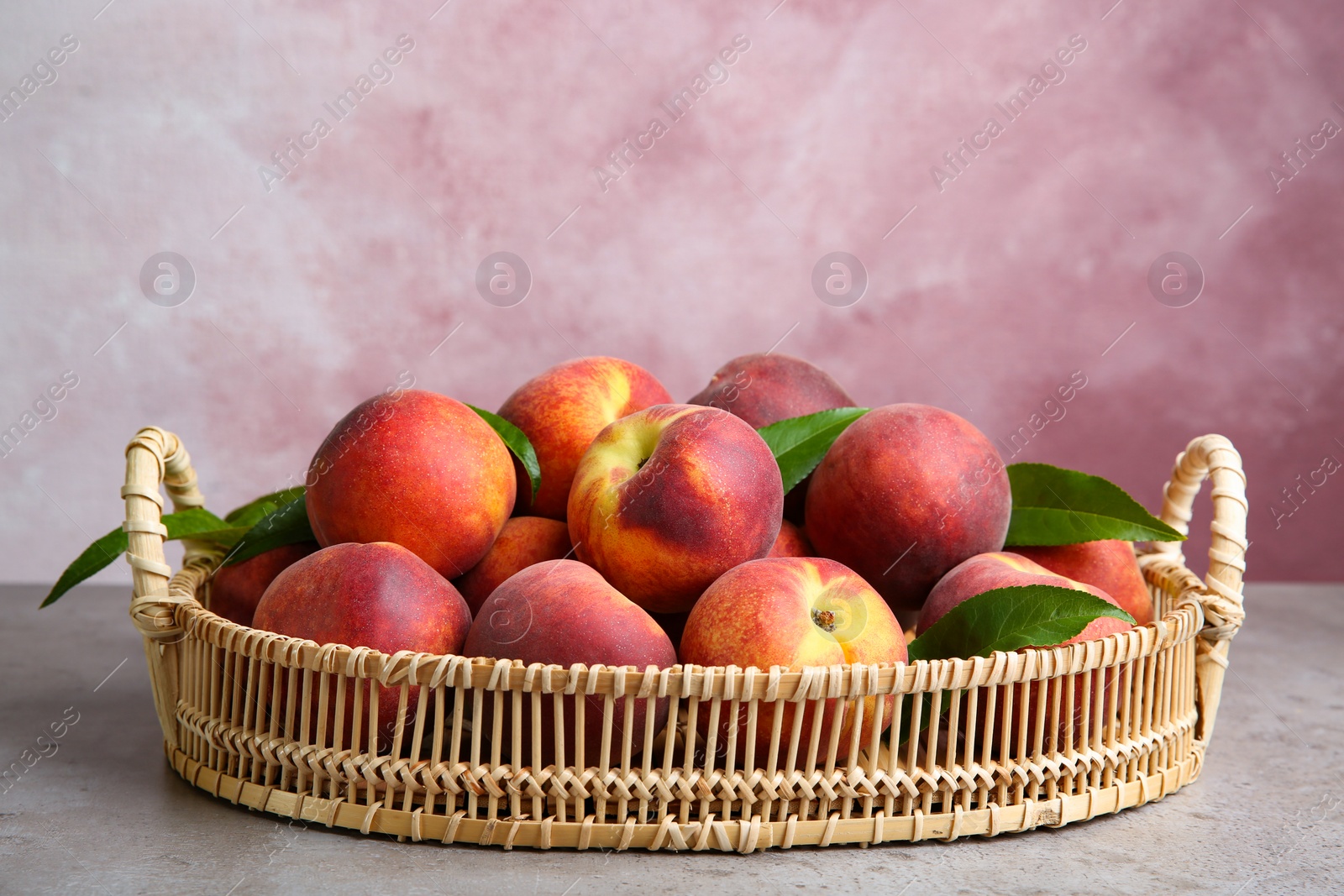 Photo of Wicker tray with tasty peaches on grey table against pink background