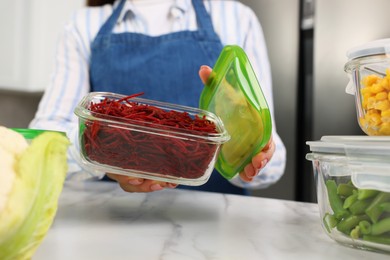 Photo of Woman holding open container with cut fresh red beets at white marble table in kitchen, closeup. Food storage
