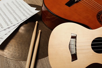 Photo of Ukulele, acoustic guitar, drumsticks, cymbal and note sheets on wooden background, closeup. Musical instruments