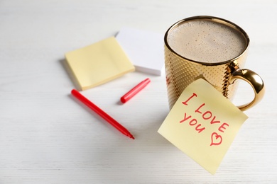 Photo of Note with phrase I Love You attached to cup of hot drink on white wooden table