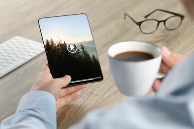 Image of Man watching video on tablet while drinking coffee at wooden desk, closeup