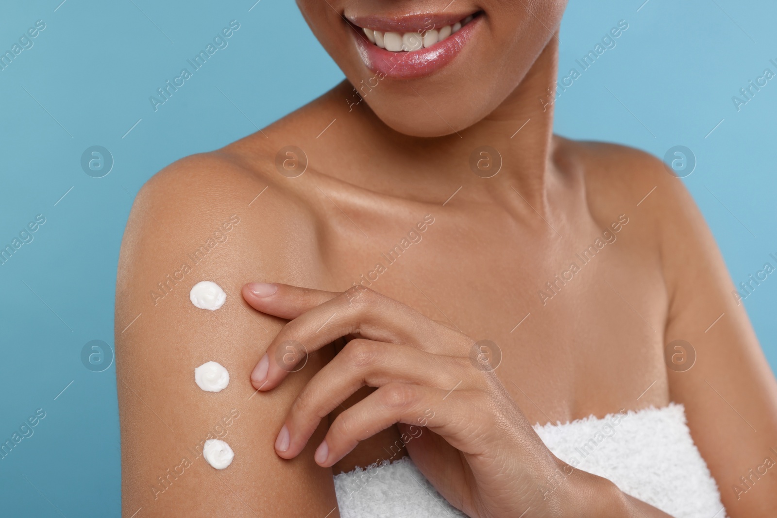 Photo of Young woman applying body cream onto arm on light blue background, closeup