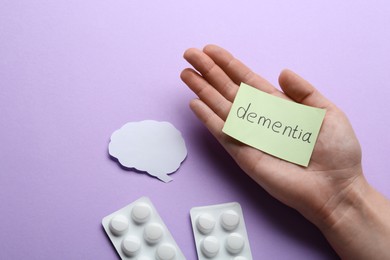 Woman holding paper note with word Dementia near pills and brain cutout on violet background, top view