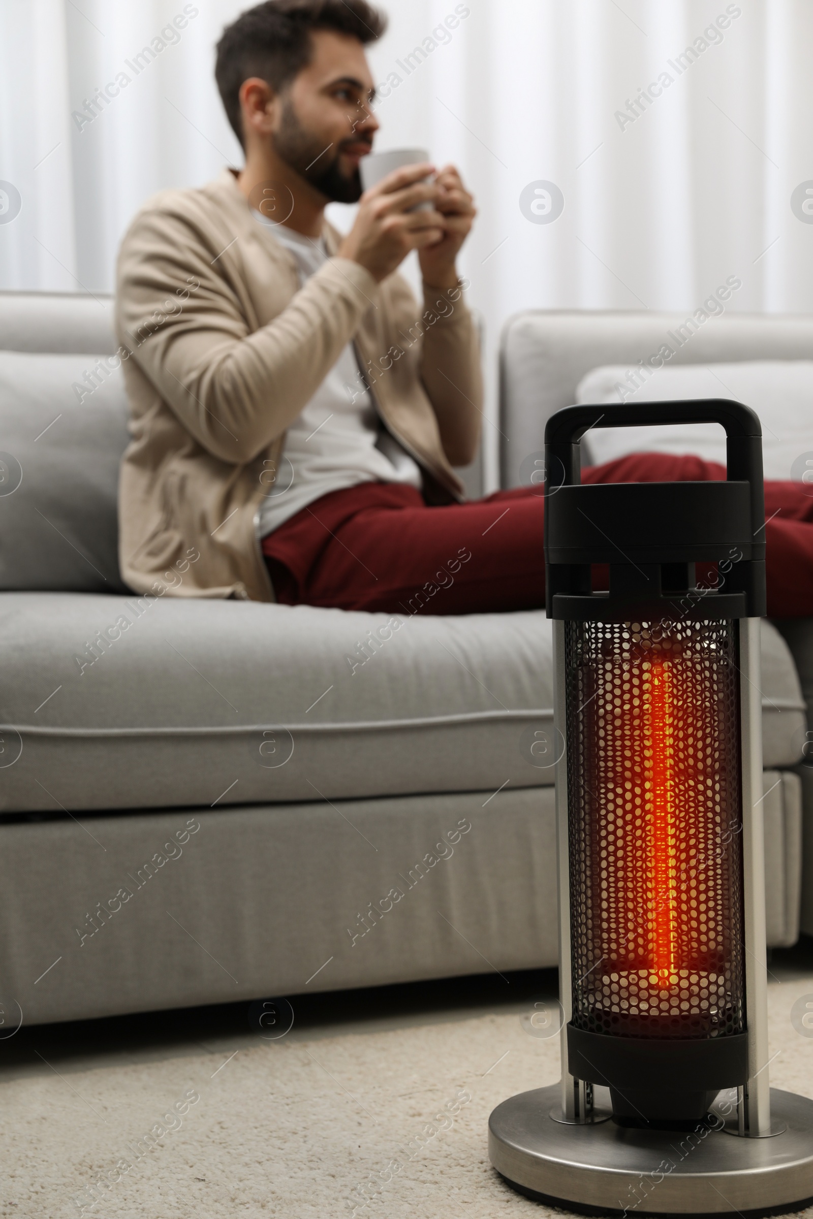 Photo of Electric heater and young man with cup of drink indoors