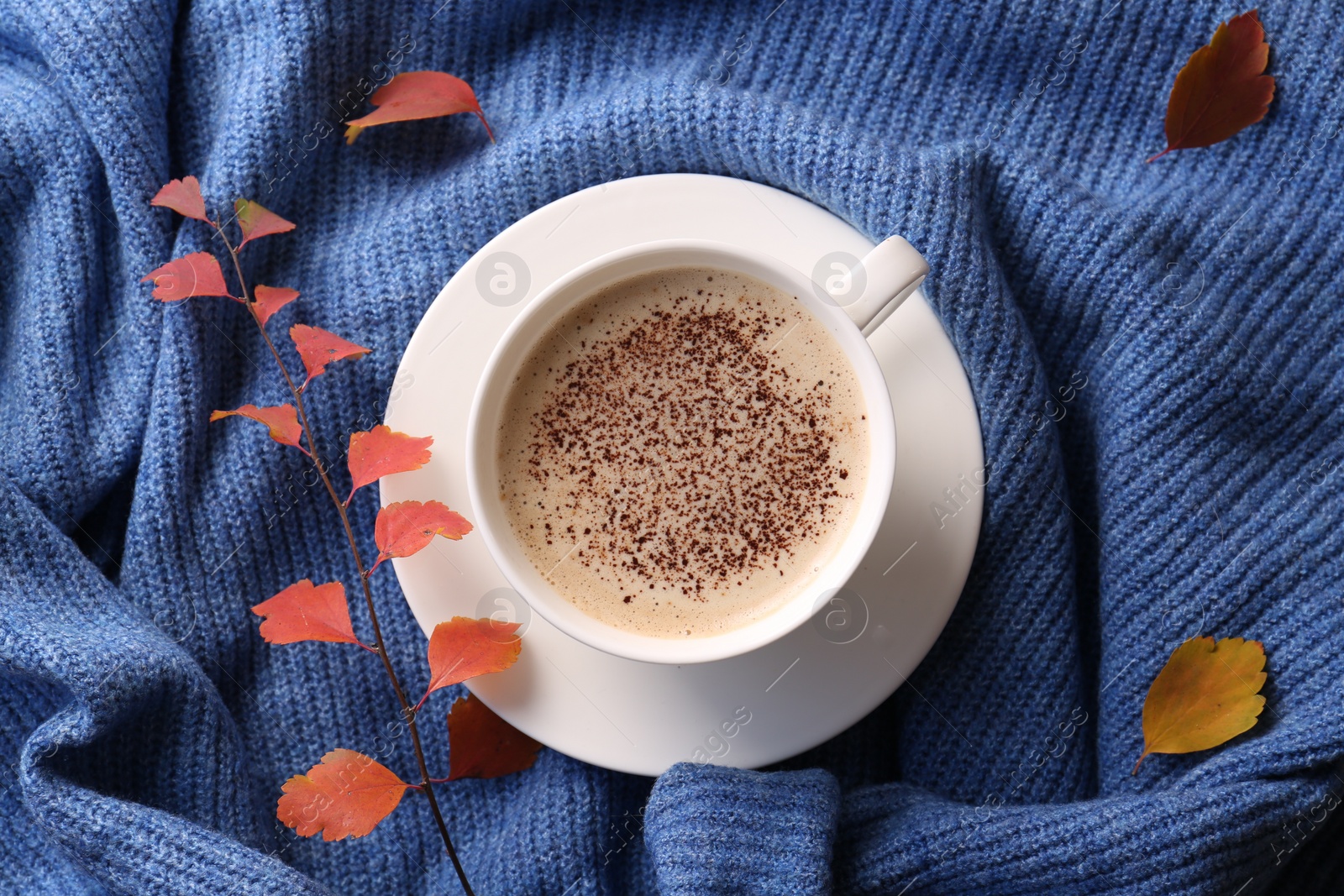Photo of Cup of hot drink and leaves on blue knitted sweater, flat lay. Cozy autumn atmosphere