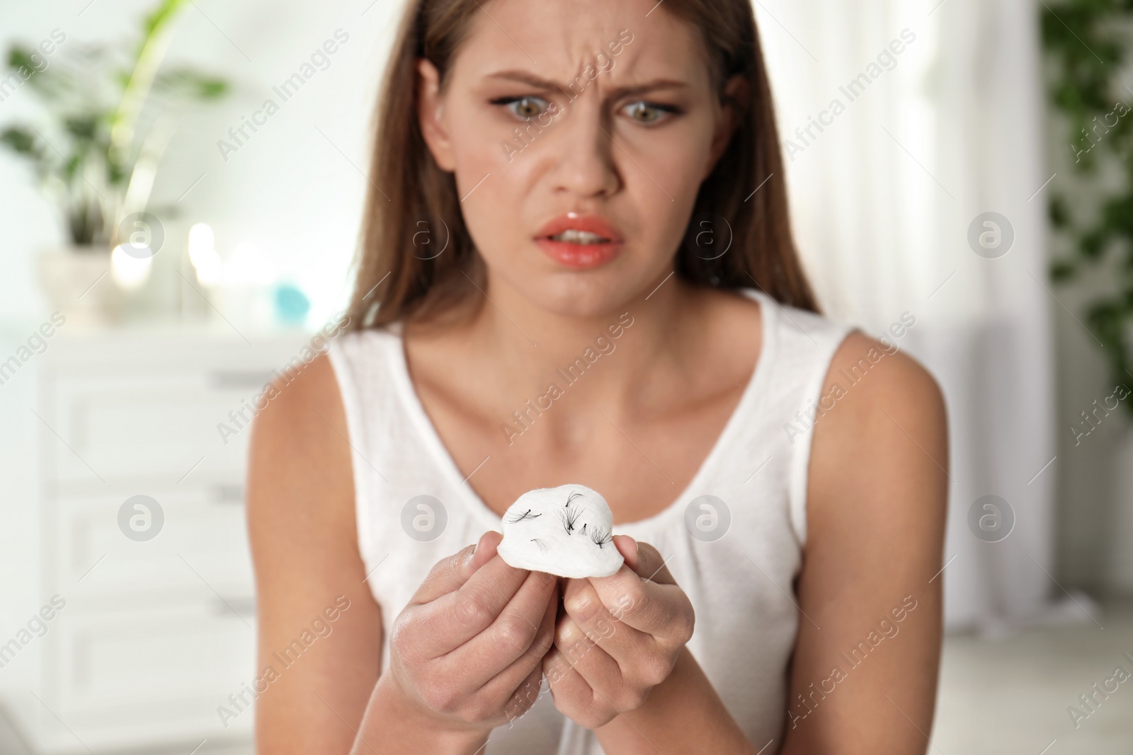 Photo of Emotional young woman holding cotton pad with fallen eyelashes indoors