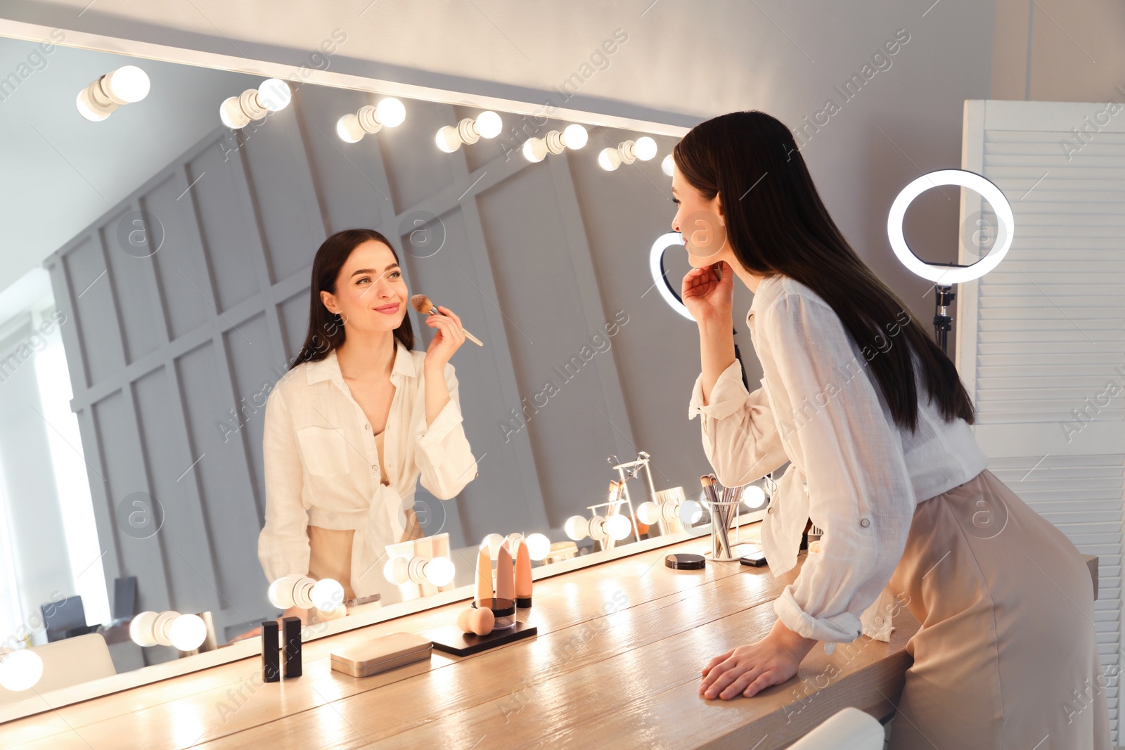 Photo of Young woman applying make up near illuminated mirror indoors