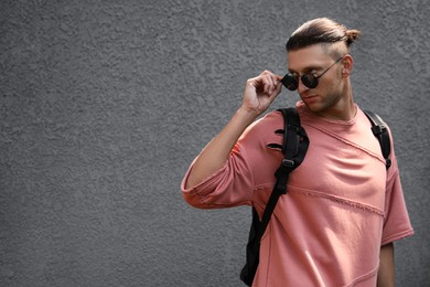 Photo of Handsome young man in stylish sunglasses and backpack near grey wall, space for text