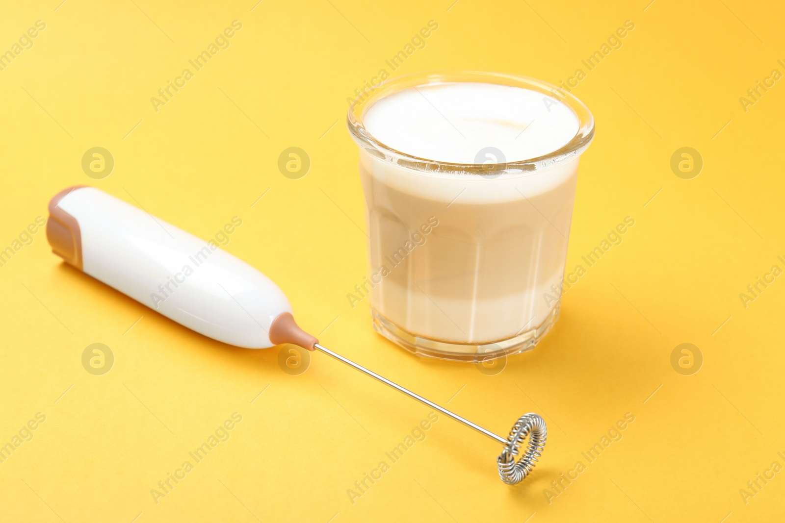 Photo of Mini mixer (milk frother) and tasty cappuccino in glass on yellow background