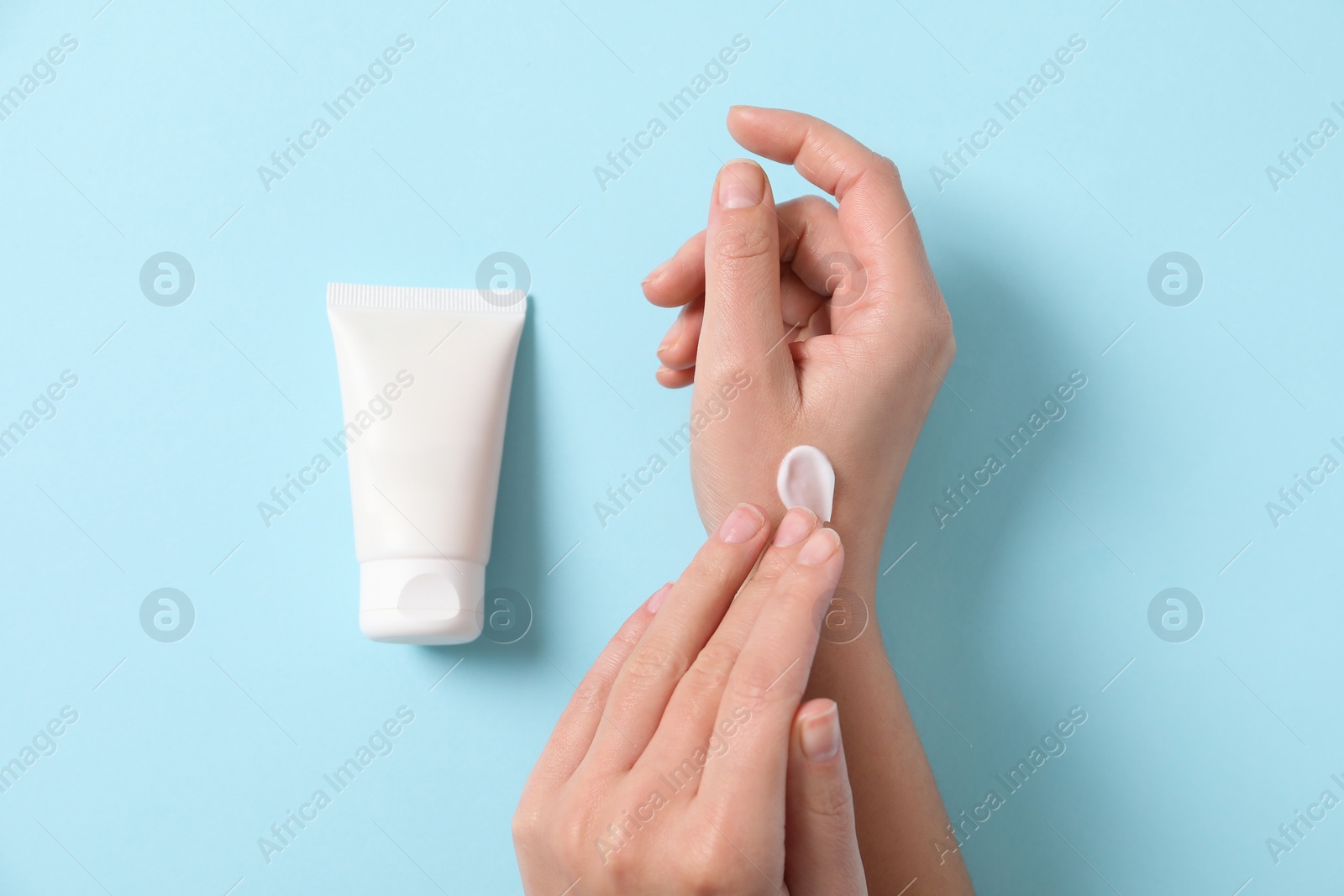 Photo of Woman with tube applying cosmetic cream onto her hand on light blue background, top view