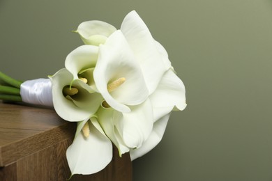 Photo of Beautiful calla lily flowers tied with ribbon on wooden table near olive wall, closeup. Space for text