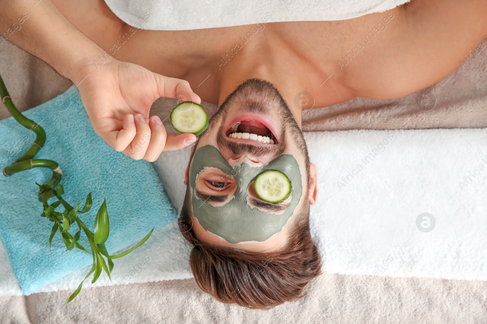 Photo of Young man with clay mask on his face holding cucumber slices in spa salon, above view