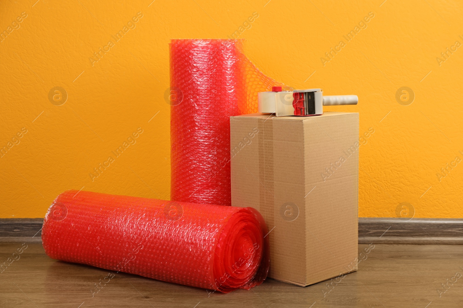 Photo of Red bubble wrap rolls, tape dispenser and cardboard box on floor near orange wall