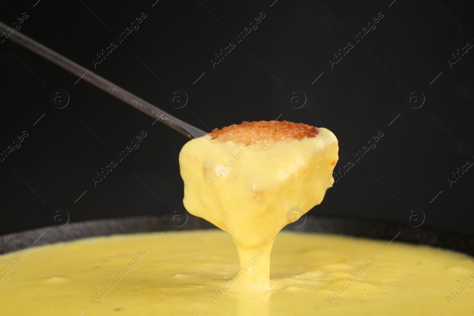 Photo of Dipping piece of bread into fondue pot with tasty melted cheese against dark gray background, closeup