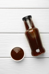 Tasty barbeque sauce in bottle and bowl on white wooden table, top view