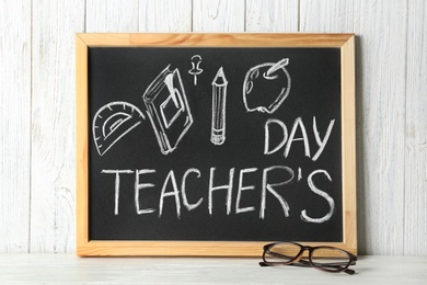 Photo of Chalkboard with inscription TEACHER'S DAY and glasses on wooden table against white wall