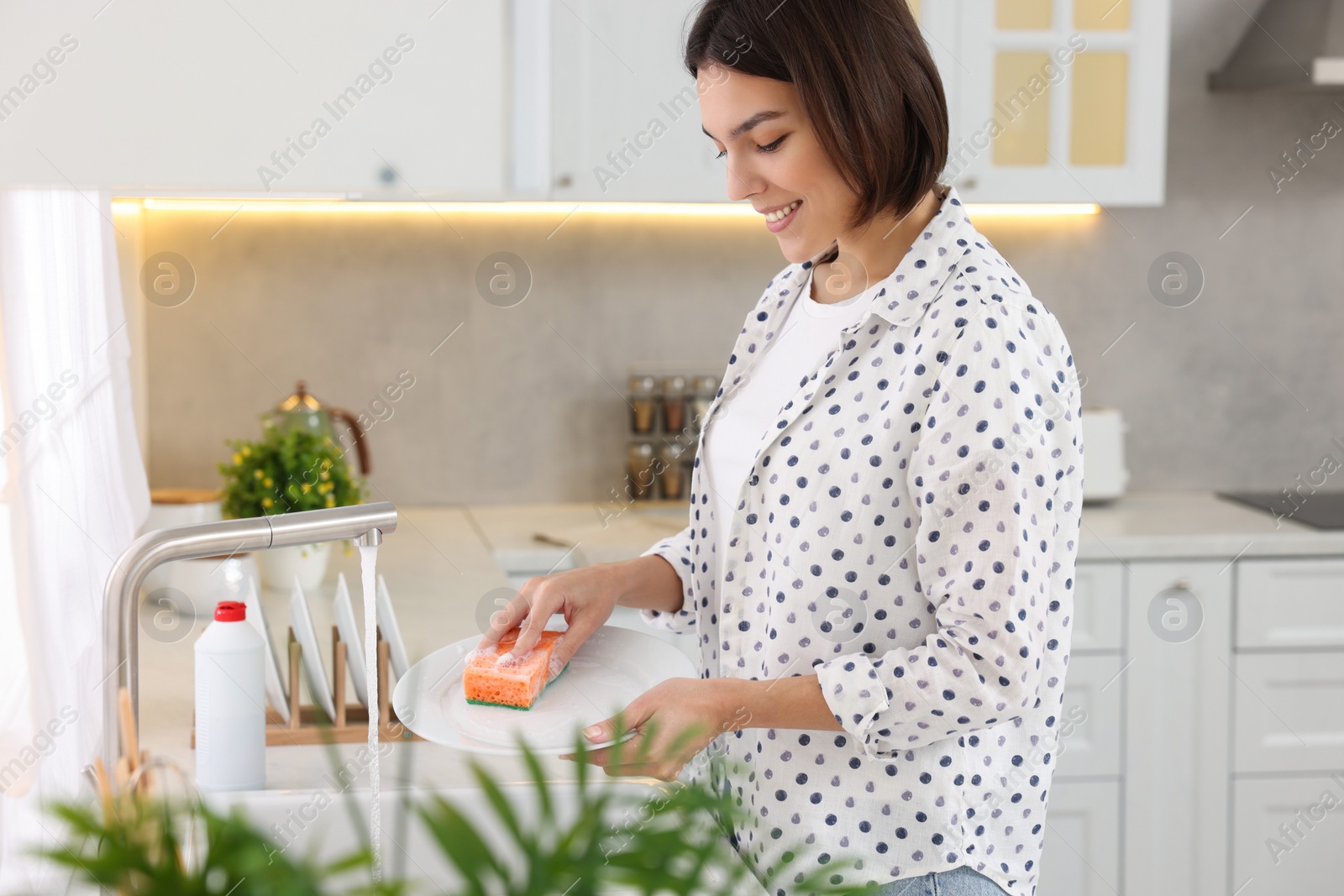 Photo of Happy young woman washing plate above sink in modern kitchen