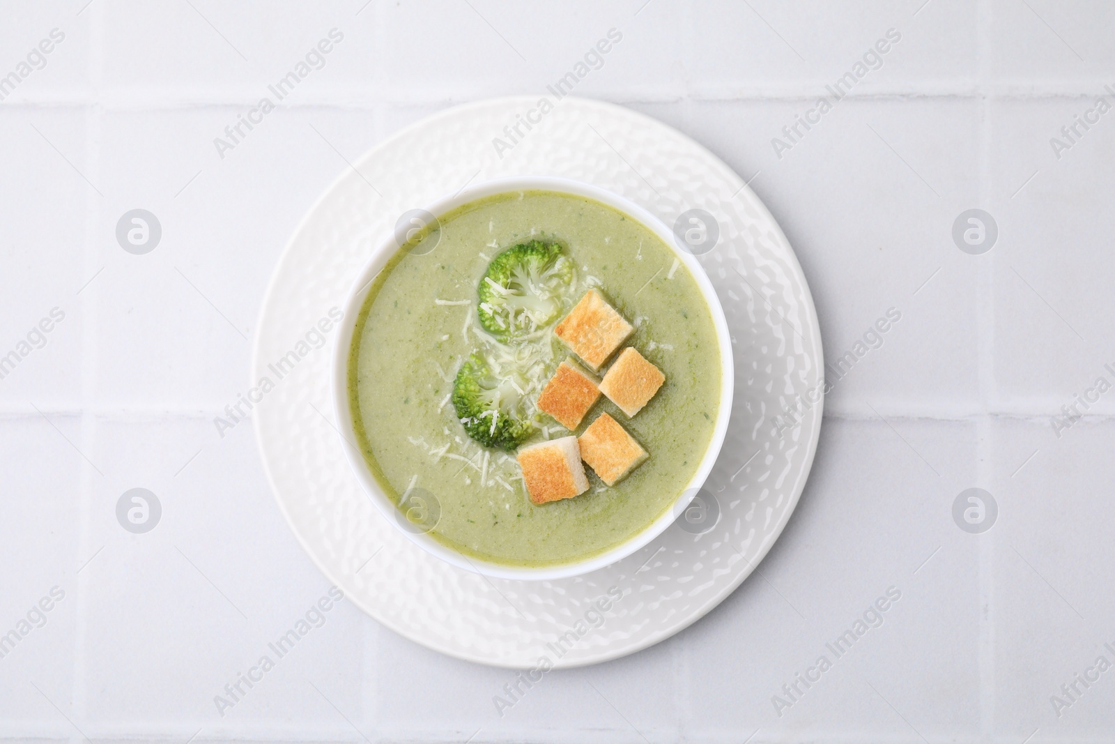 Photo of Delicious broccoli cream soup with croutons and cheese on white tiled table, top view