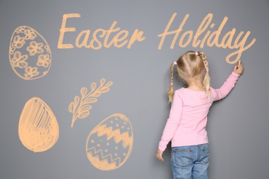 Image of Small girl writing phrase Easter Holiday on grey wall with drawings. School holiday
