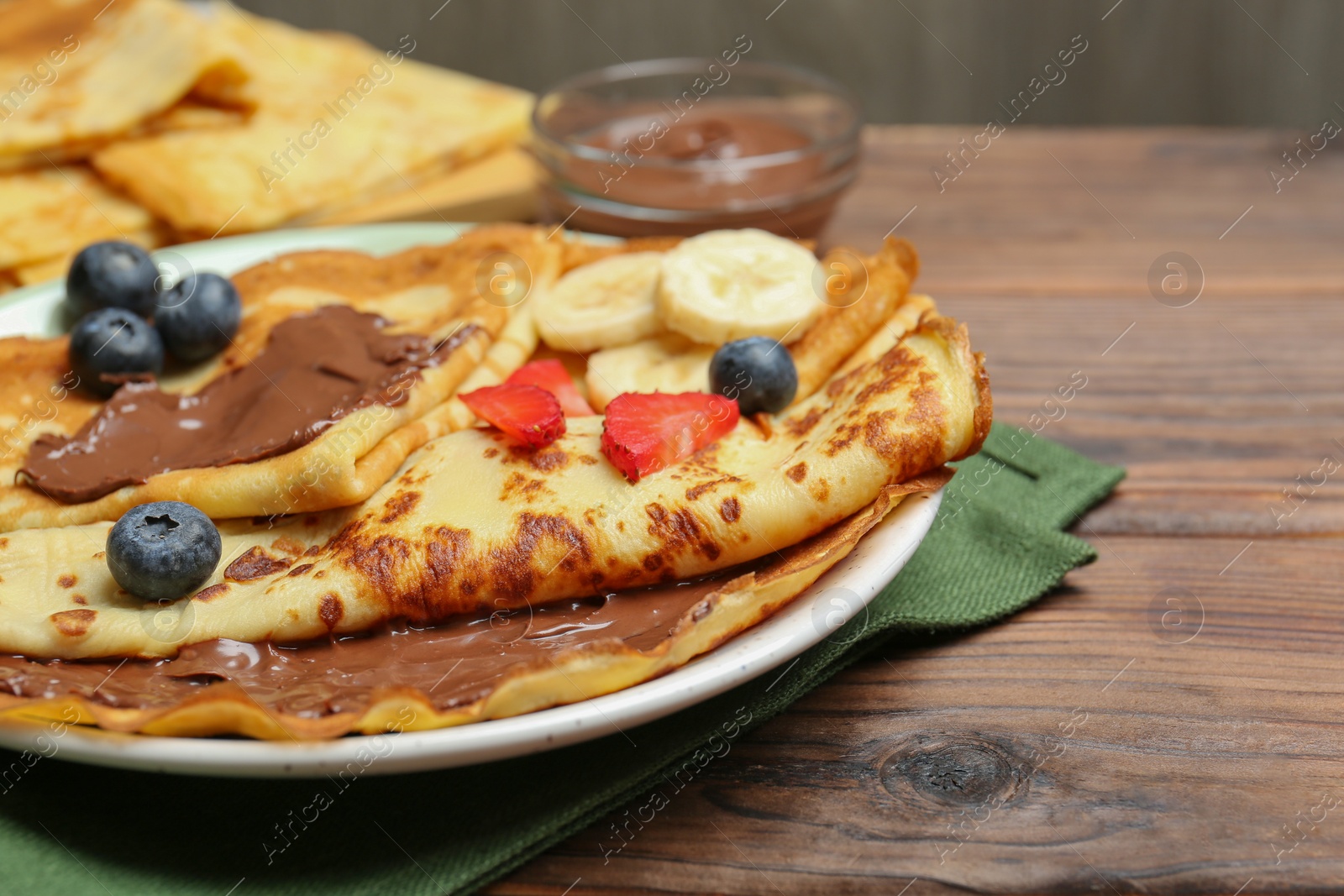 Photo of Tasty crepes with chocolate paste, banana and berries on wooden table, closeup