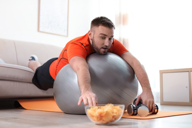 Photo of Lazy young man on exercise ball reaching for chips at home