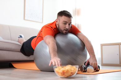 Lazy young man on exercise ball reaching for chips at home