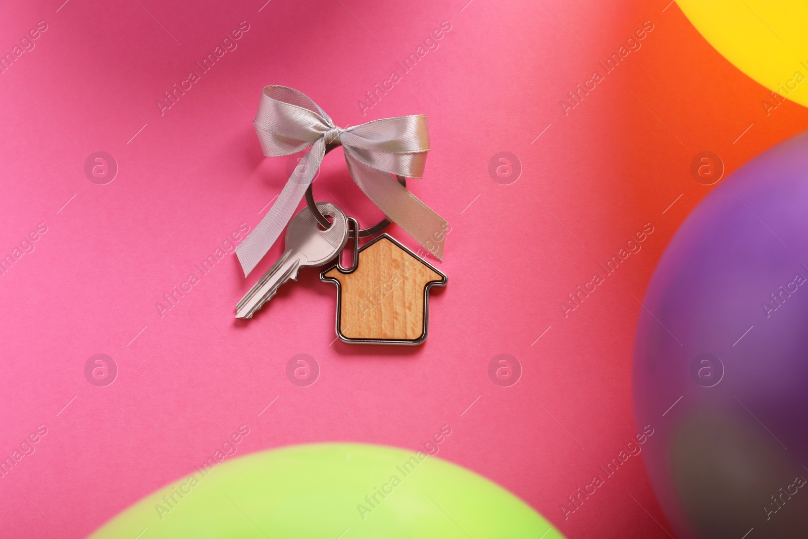 Photo of Key with trinket in shape of house and bow on pink background near color balloons, flat lay. Housewarming party