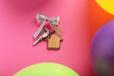 Key with trinket in shape of house and bow on pink background near color balloons, flat lay. Housewarming party