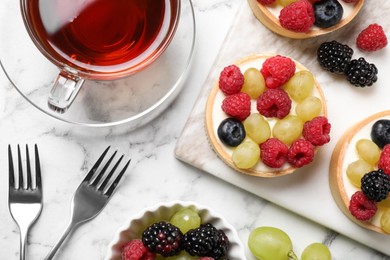 Delicious tartlets with berries and tea on white marble table, flat lay