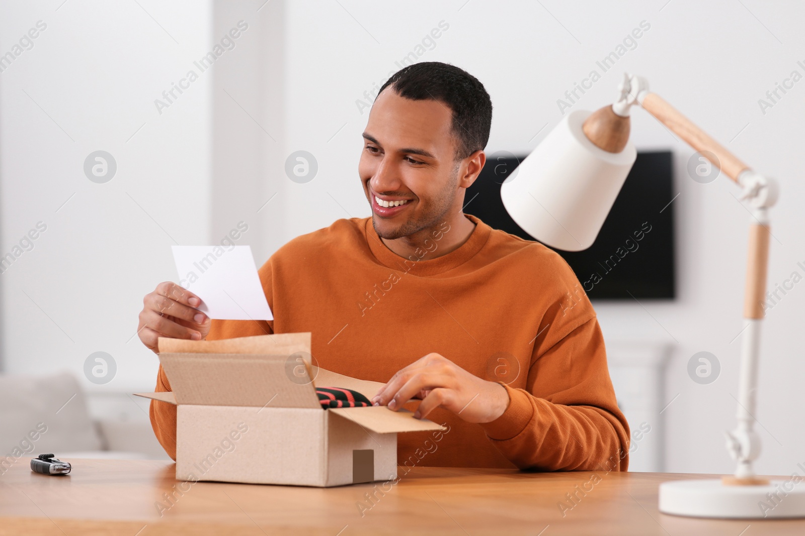 Photo of Happy young man with greeting card near parcel at wooden table. Internet shopping