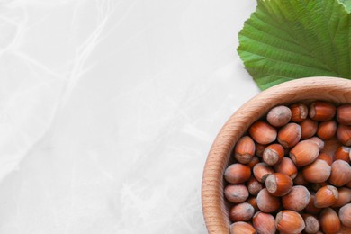 Photo of Tasty hazelnuts in wooden bowl and leaf on light marble table, flat lay. Space for text
