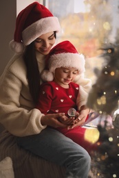 Photo of Mother and daughter in Santa hats playing with snow globe near window