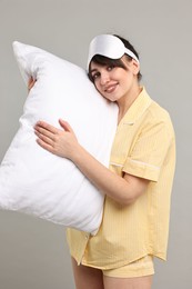 Happy woman in pyjama and sleep mask holding pillow on grey background